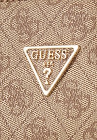 Tissu marron texturé avec un motif répété du logo GUESS et un emblème métallique triangulaire doré GUESS USA avec un point d'interrogation au centre.