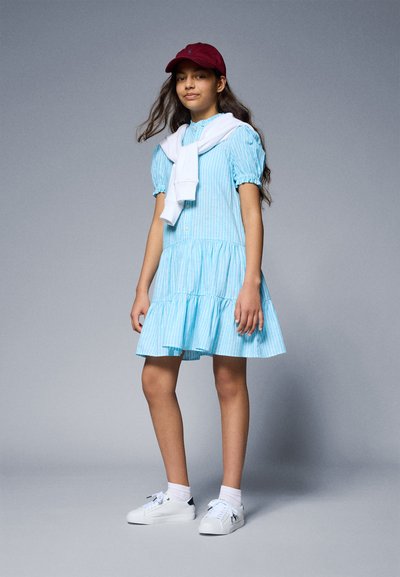 Robe rayée bleu clair avec des manches bouffantes et une jupe à volants, associée à un pull blanc enroulé autour du cou et une casquette bordeaux. Baskets blanches.