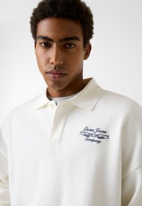 White polo shirt featuring a collared design, button placket, and blue embroidered logo with "Guess Jeans Company" and "EST. 1981."