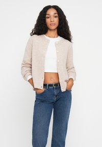 Light beige knitted cardigan with round neckline, button closure, and ribbed cuffs, paired with high-waisted blue jeans.