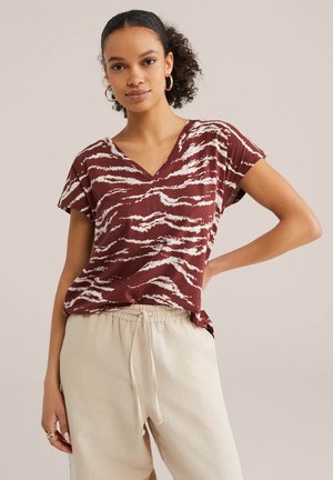 Vrouw met krullend haar, goudkleurige oorringen, een bordeauxrode en witte gestreepte V-hals top en beige broek met trekkoord, staand met één hand op haar heup.