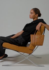 Woman in black outfit reclining on tan leather chair, holding a brown patterned handbag with gold accents.