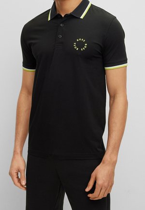 Polo noir en coton, avec un logo jaune sur la poitrine et des accents verts contrastants sur le col et les manches.