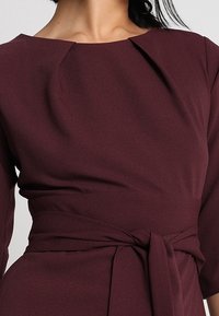 Robe bordeaux avec un décolleté bateau, des manches longues et un détail de ceinture nouée. Tissu lisse avec un léger drapé aux épaules.