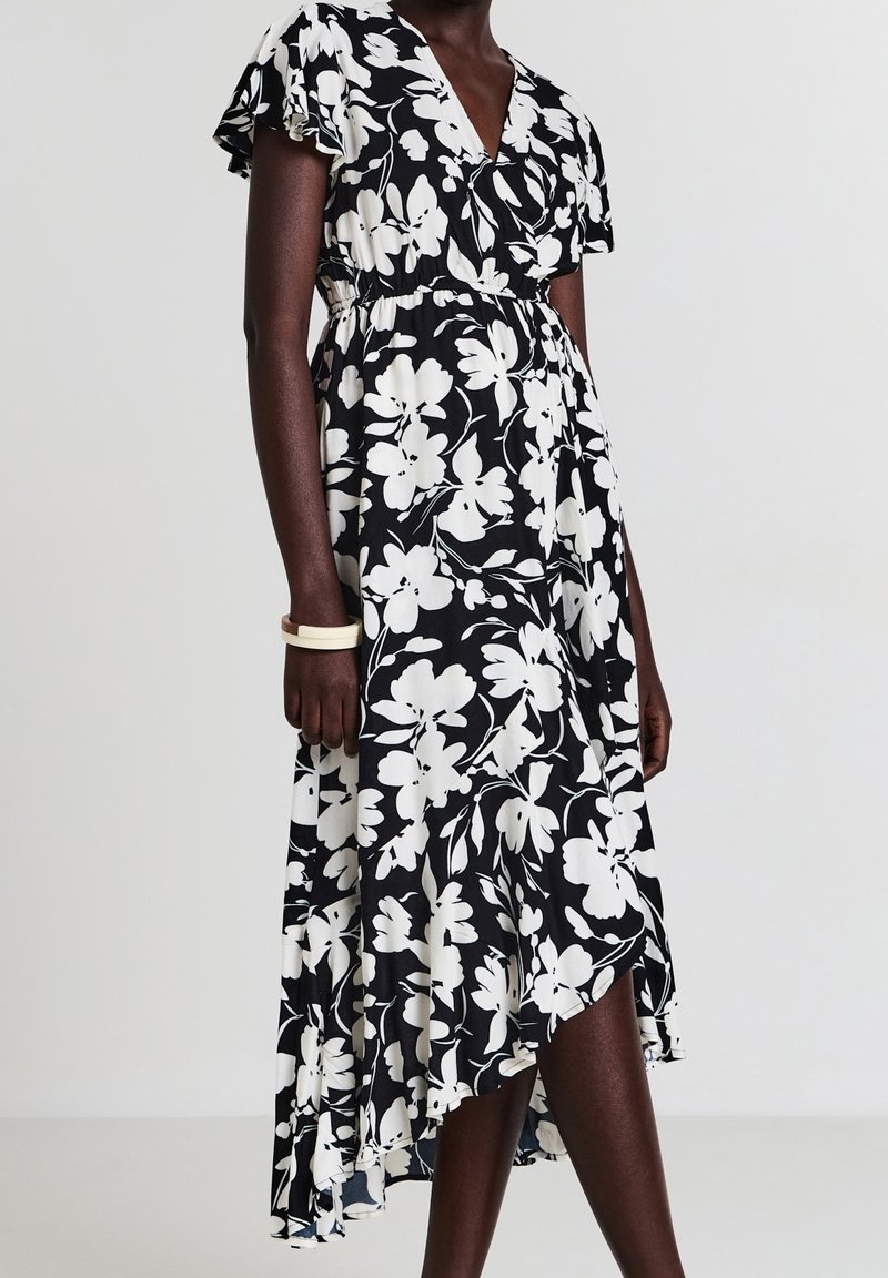 Femme portant une robe portefeuille fleurie noire et blanche avec des manches volantées et un ourlet asymétrique, accessoirisée d'un bracelet blanc.