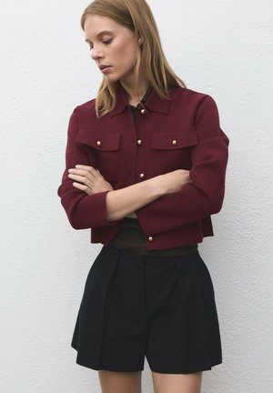 Bordeaux cropped jack met gouden knoopaccenten, twee voorzakken, gecombineerd met zwarte high-waisted shorts, gedragen over een aansluitend topje.