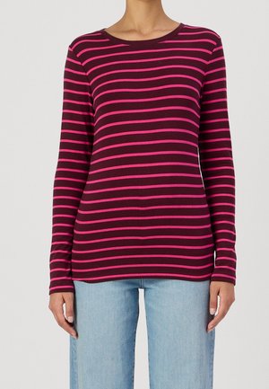 Personne portant une chemise à manches longues rayée horizontalement rouge foncé et rose associée à un jean en denim bleu clair, debout avec les bras détendus.