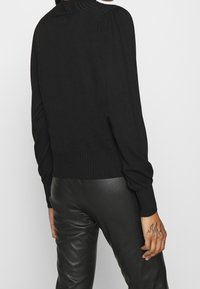 Schwarzer Strickpullover mit hohem Rippkragen, überschnittenen Schultern und gerippten Bündchen. Kombiniert mit engen schwarzen Lederhosen.