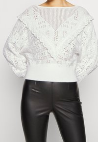 Pull en laine blanc avec un col festonné, présentant un motif texturé. L'ourlet côtelé repose au-dessus d'un legging en cuir noir.