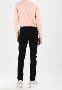 Svarta jeans med slim fit, tillverkade av denim. Bärs med en ljusrosa långärmad skjorta och grå sneakers, sedd bakifrån.