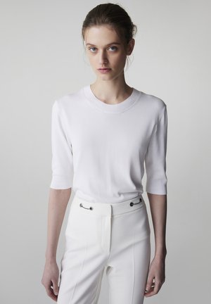 Jeune femme aux cheveux clairs tirés en arrière, portant un haut blanc à manches courtes et un pantalon blanc ajusté avec des détails de boutons métalliques.