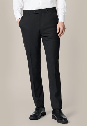 Mężczyzna ubrany w dopasowane czarne spodnie garniturowe, czarne skórzane buty typu brogue i białą koszulę zapinaną na guziki, z rękami w kieszeniach.