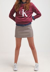 Sudadera burdeos de Calvin Klein con logotipo blanco, superpuesta sobre una camisa de mezclilla azul. Falda mini gris, zapatillas blancas, en posición de pie, fondo liso.