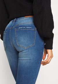 Jeanși subțiri din denim albastru, cu talie înaltă, buzunare pe spate și detalii ușor distresate, asortați cu un pulover tricotat negru.