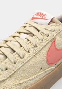 Beige textured sneaker with a gum sole, pink Nike swoosh, and off-white laces. Features a rubberized tongue and stitched detailing.