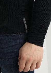 Czarny sweter z dzianiny z ribbami, z boczną metką oznaczoną "TOM TAILOR ACTIVE." Ręka oparta na talii, obok ciemnych dżinsów.