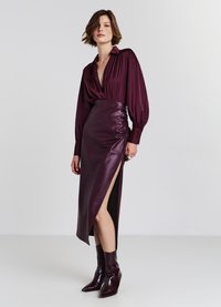 Blusa di seta bordeaux con scollatura a V profonda e maniche lunghe, abbinata a una gonna in pelle aderente con uno spacco laterale e stivali al ginocchio.