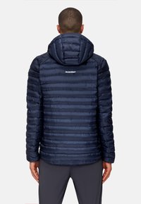 Marineblå pufferjakke med hætte, der har et vandret quiltet design. "Mammut" logo på den øverste ryg. Glat, skinnende overflade.