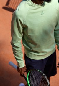 Felpa verde chiaro con maniche raglan, abbinata a pantaloni corti scuri. L'individuo tiene in mano una racchetta da tennis con un'impugnatura verde.