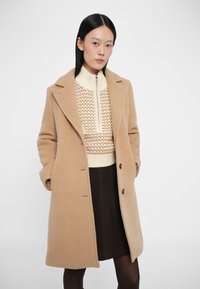 Abrigo de mezcla de lana beige con cuello de pico y botones frontales, superpuesto a un suéter de punto crema con patrones y una falda marrón oscuro.