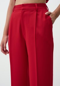 Red tailored trousers with a smooth texture, featuring front pleats, side pockets, and a fitted waistband.