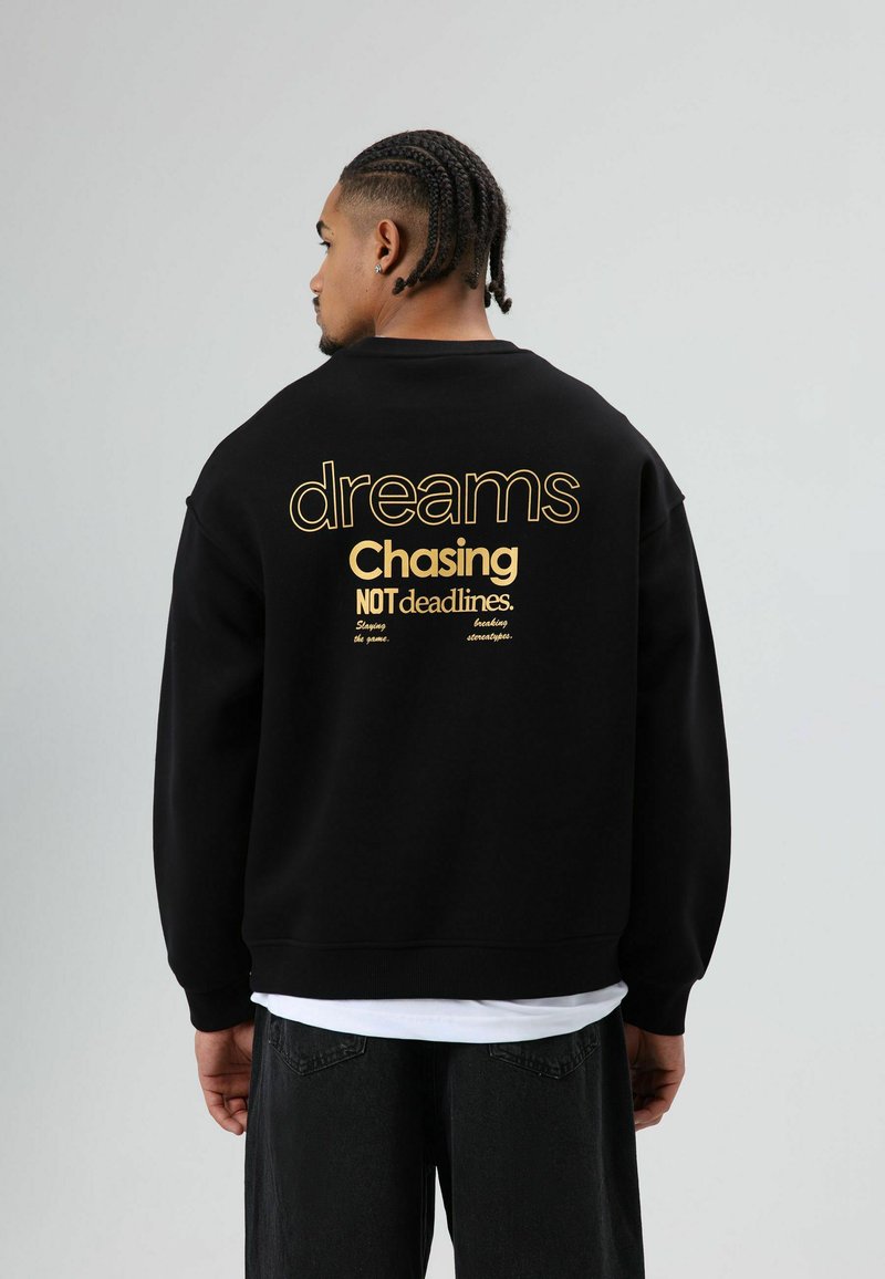 Zwarte sweatshirt met gouden tekst op de achterkant met de woorden "dreams Chasing NOT deadlines." Voorzien van geribde manchetten en een ontspannen pasvorm.