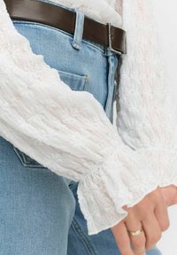 Vit, texturerad blus med volanger vid ärmarna som är ihopparad med ljusblå denimjeans. Ett brunt läderbälte är synligt vid midjan.