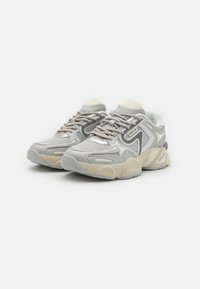 Des baskets en maille grises présentent un design superposé avec des accents beiges, des éléments réfléchissants et des surfaces texturées sur une semelle épaisse.