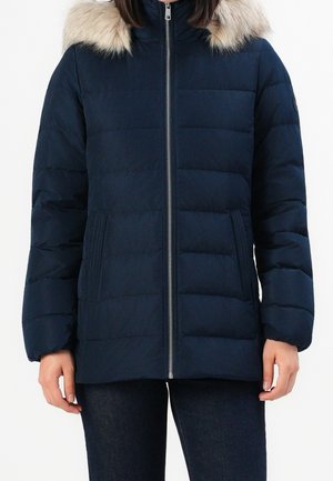 Marineblaue gesteppte Winterjacke mit silbernem Reißverschluss und beigem Kunstpelzbesatz an der Kapuze, getragen mit dunklen Jeans.