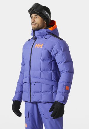 Hombre con chaqueta acolchada morada y pantalones con acentos naranjas, guantes negros, gorro negro y gafas de esquí en la frente, mirando hacia la izquierda.