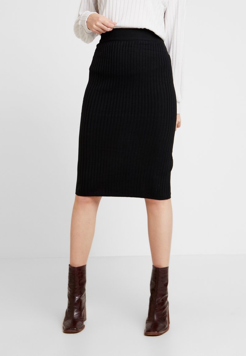 Schwarzer gerippter Strickrock mit hohem Bund, knielangem Design, strukturiertem Stoff und figurbetontem Schnitt. Getragen mit braunen Ankle Boots.