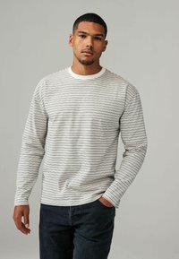 Langærmet stribet skjorte i creme og marineblå, med en rund halsudskæring og tekstureret stof. Båret med mørke jeans, hænderne i lommerne.