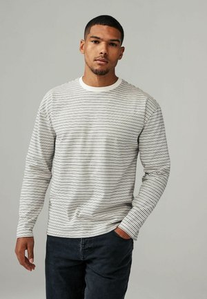 Langærmet stribet skjorte i creme og marineblå, med en rund halsudskæring og tekstureret stof. Båret med mørke jeans, hænderne i lommerne.