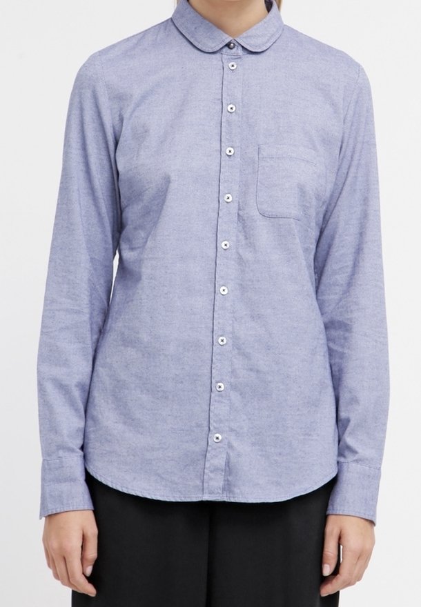 Femme portant une chemise bleu clair à manches longues boutonnée avec un col arrondi et une poche poitrine, associée à un pantalon noir.