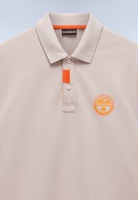 Beige polo skjorte med en blød tekstur, prydet med en orange detalje i kraven og et rundt logo i orange og hvidt på brystet.