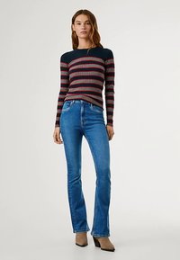 Stripete ribbestrikket genser i marineblå, burgunder og grå. Figurformet design. Kombinert med lyseblå vide jeans og mørke sko med hæl.