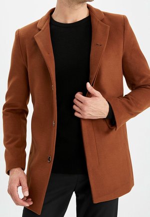 Mężczyzna poprawiający zapięty brązowy wełniany płaszcz noszony na czarnym swetrze i czarnych spodniach, na tle jasnego, jednolitego tła.