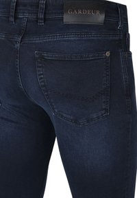 Dark blue denim jeans with a fitted design, featuring a back pocket, stitched detailing, and a label with "GARDEUR" near the waistband.