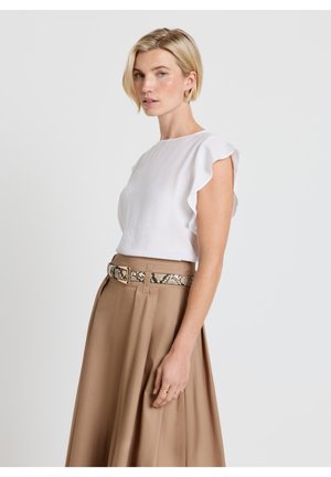 Frau mit kurzen blonden Haaren, die ein weißes ärmelloses Oberteil mit Rüschen und einen hoch taillierten, hellbraunen Rock mit Schlangenmuster-Gürtel trägt, seitlich stehend.