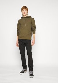 Felpa verde oliva con tasca frontale e cappuccio regolabile, abbinata a pantaloni neri e scarpe Vans nere con un motivo di logo bianco.