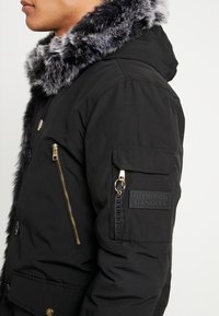 Personne portant une veste d'hiver noire avec col en fausse fourrure grise, fermeture éclair dorée sur la poche poitrine et écusson logo Glorious Gangsta sur la poche de la manche.