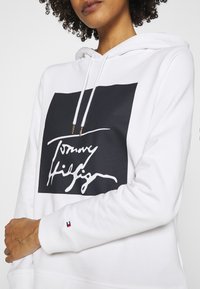 Sudadera blanca con un gráfico cuadrado negro que presenta "Tommy Hilfiger" en cursiva blanca. Cordones con puntas doradas y un pequeño logo en el puño.