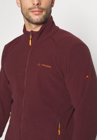 Giacca in pile rosso scuro con colletto alto, chiusura frontale con zip e dettagli arancioni. Presenta due tasche laterali con zip e dettagli del logo.
