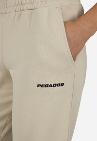 Bézs színű, puha anyagból készült melegítő nadrág, rugalmas derékrésszel és oldalzsebekkel; oldalán fekete "PEGADOR" logó hímzéssel.
