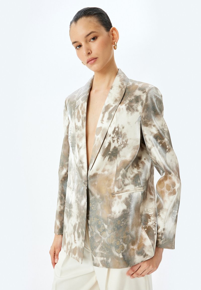 Blazer i en beige, grå og creme tie-dye mønster. Har en enkelt knap, spidse rever og to forlommer. Glat, skinnende stof.