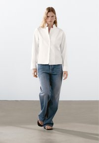 White button-up shirt with a collar, paired with loose blue denim jeans and black sandals. Clean lines, simple design, and tailored fit.