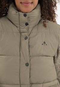 Beige Steppjacke mit hohem Kragen, ausgestattet mit schwarzen Knöpfen und einem kleinen Logo im oberen linken Bereich. Sichtbares strukturiertes Gewebe.