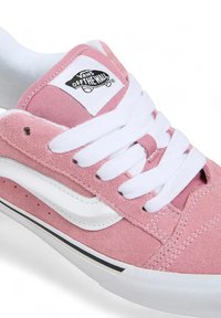 Le sneakers Vans rosa chiaro presentano una tomaia in suede, lacci bianchi e una striscia laterale caratteristica. Includono un logo in bianco e nero sulla linguetta.