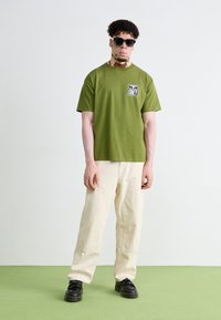 T-shirt vert olive avec un logo sur le devant, coupe décontractée. Associé à un pantalon cargo crème et des chaussures noires, sur un fond clair avec un sol vert.