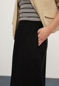 Crne hlače s elastičnim pojasom i bočnim džepovima, uparen s prugastom crno-bijelom majicom i bež jaknom s patentnim zatvaračem.